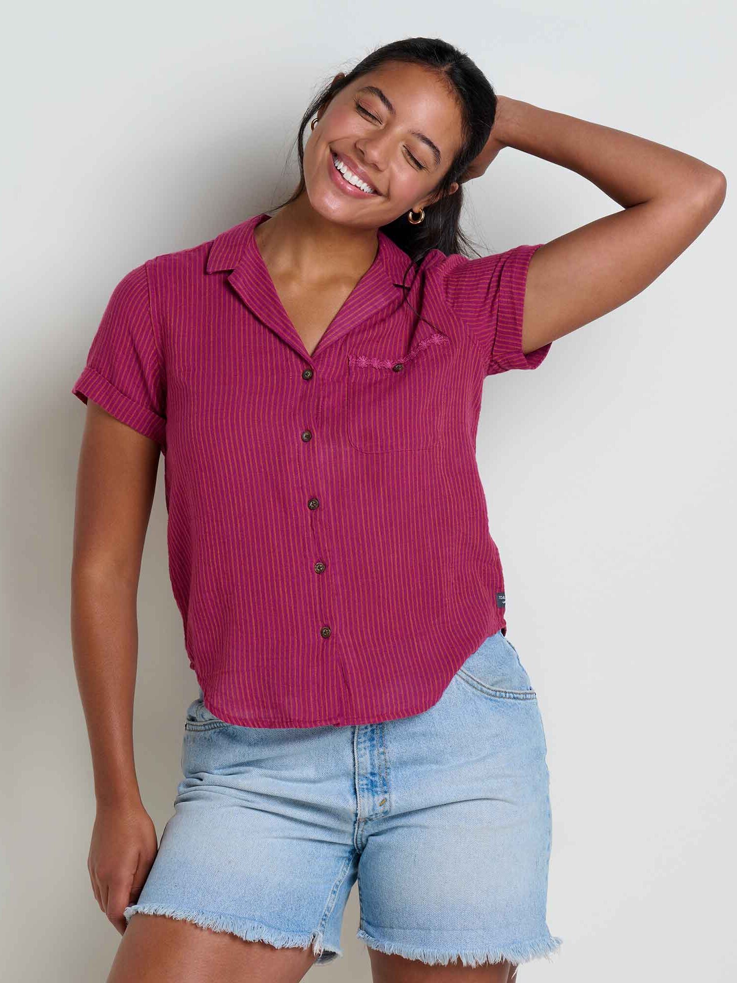 A woman wearing a short-sleeved, button-up magenta shirt and light denim shorts stands against a plain background, smiling with one hand behind her head.