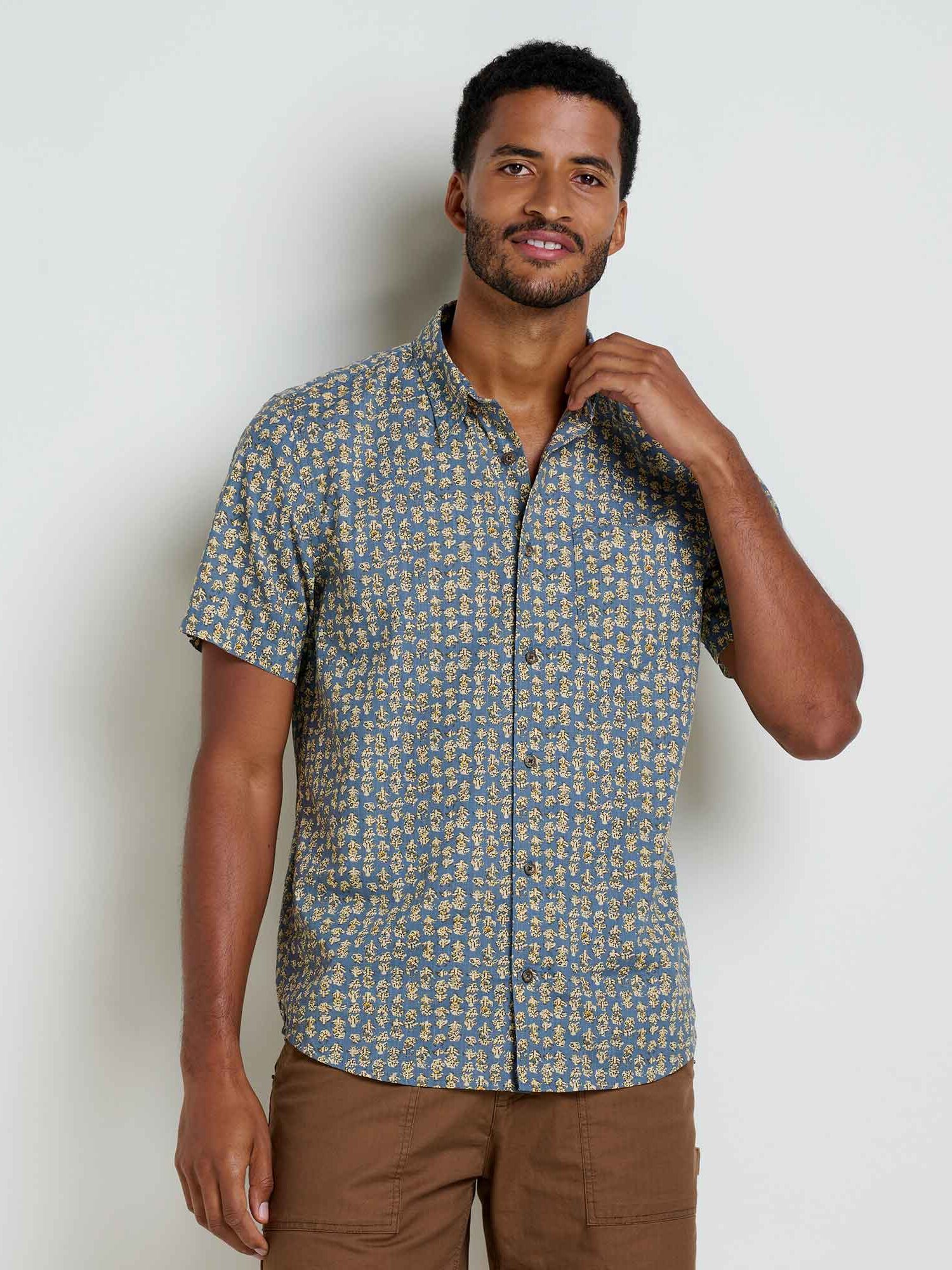 A man stands against a plain light-colored wall wearing a short-sleeve, button-up shirt with a small floral print and light brown pants.