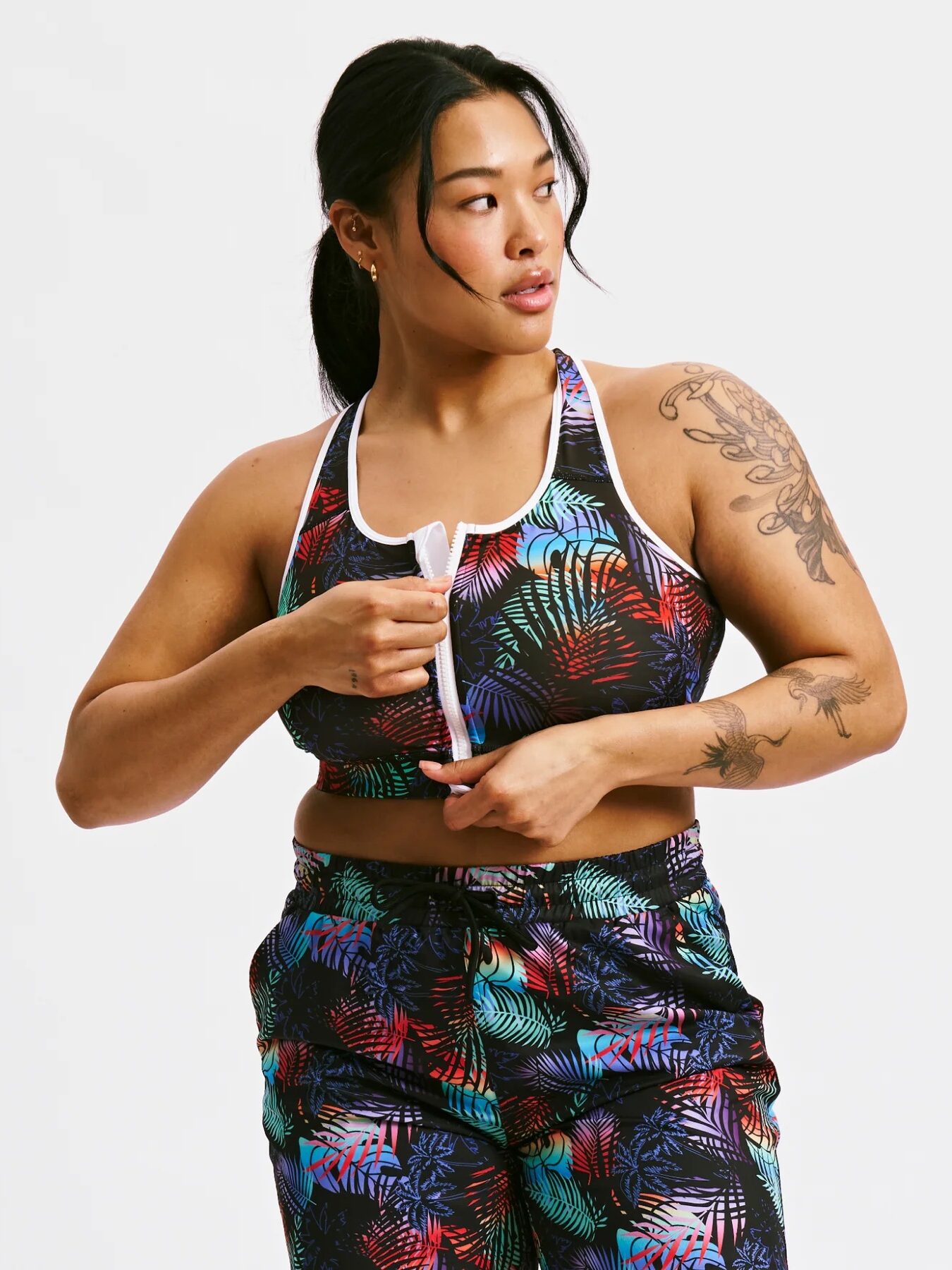 Woman in a colorful tropical-patterned sports bra and pants zipping up the front of her top, looking to her left against a plain white background.