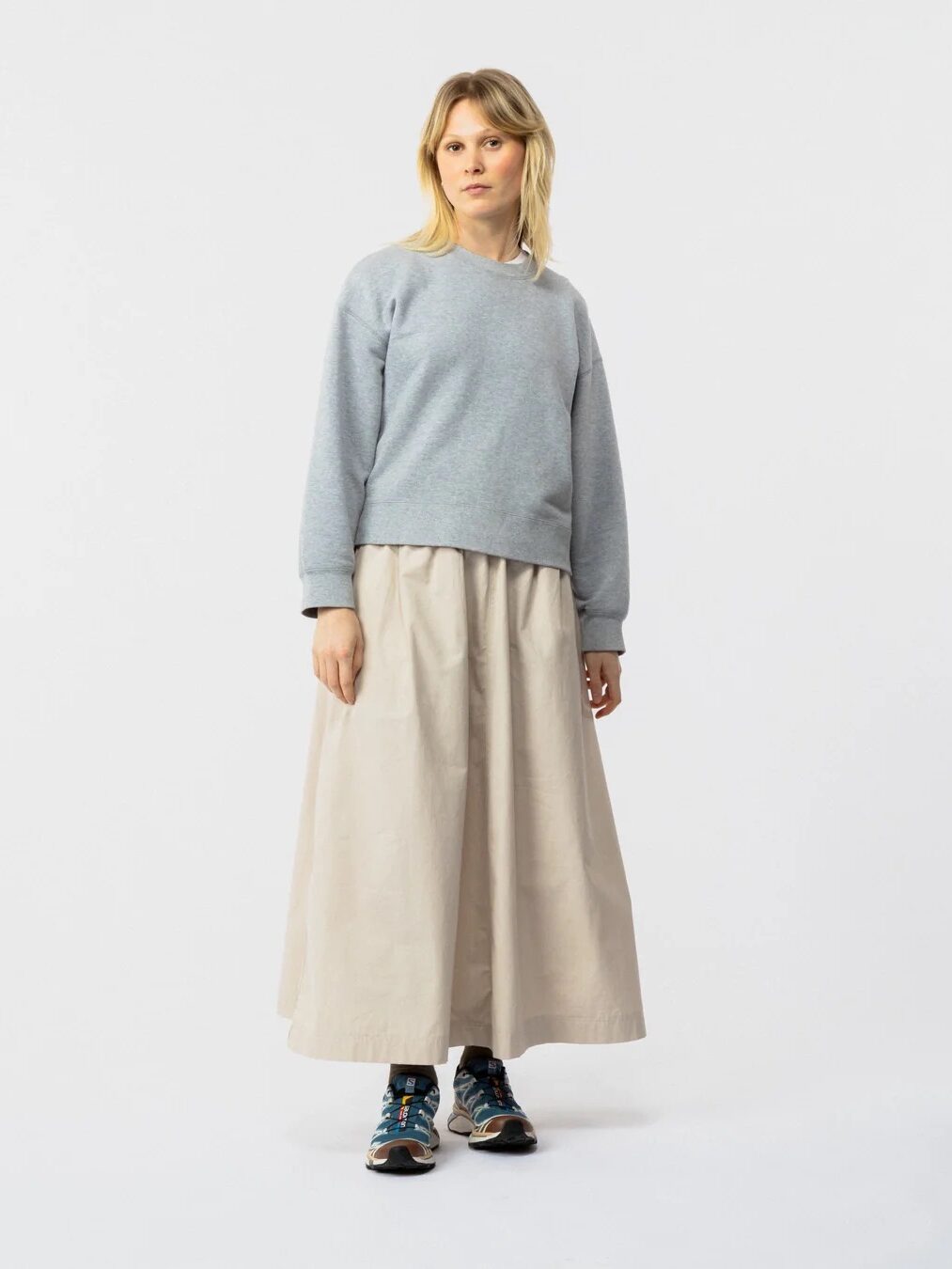 A person stands against a plain background wearing a light gray sweatshirt, a long beige skirt, patterned socks, and black sneakers.