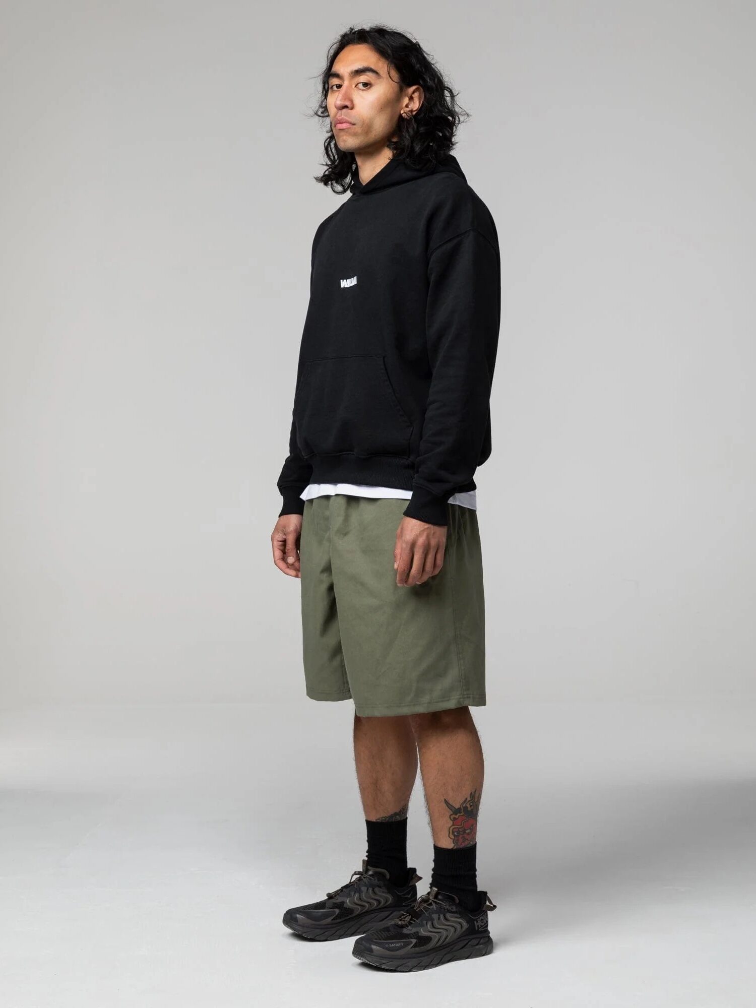 A person stands in a studio wearing a black hoodie, olive green shorts, black socks, and black sneakers, with a tattoo visible on their left leg.