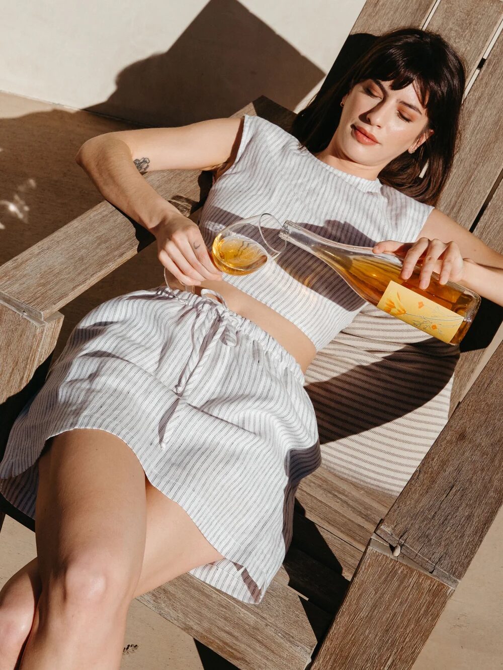 A woman in a striped crop top and skirt lounges on a wooden chair outdoors, pouring a drink from a bottle into a glass.
