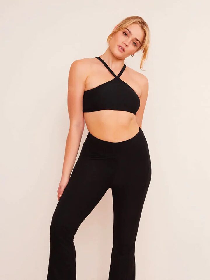 A person stands indoors on a wooden floor wearing a black crisscross crop top, black flared pants, and white sneakers, facing the camera with a neutral expression.