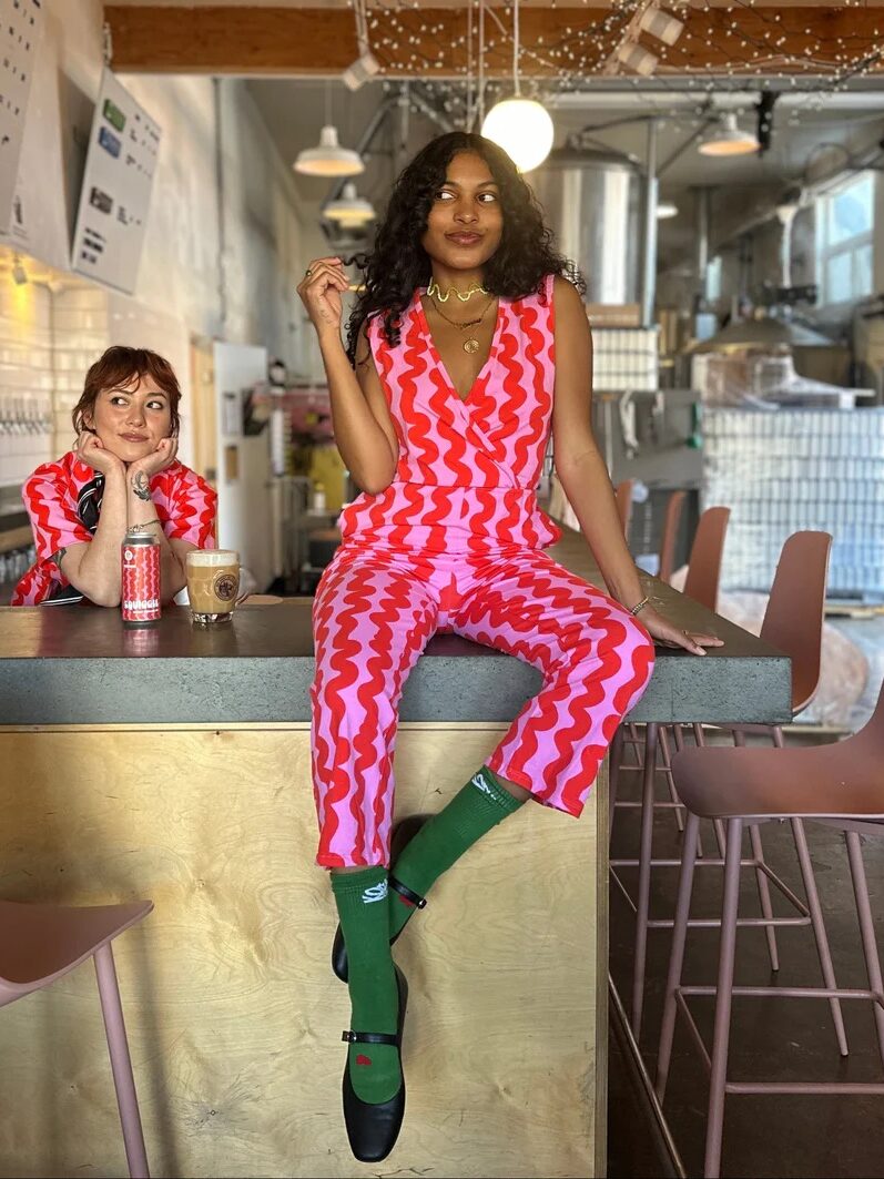 Two women in a modern cafe, one sitting on the counter in a red and pink patterned outfit with green socks, the other behind the counter wearing a red print shirt, both looking relaxed.