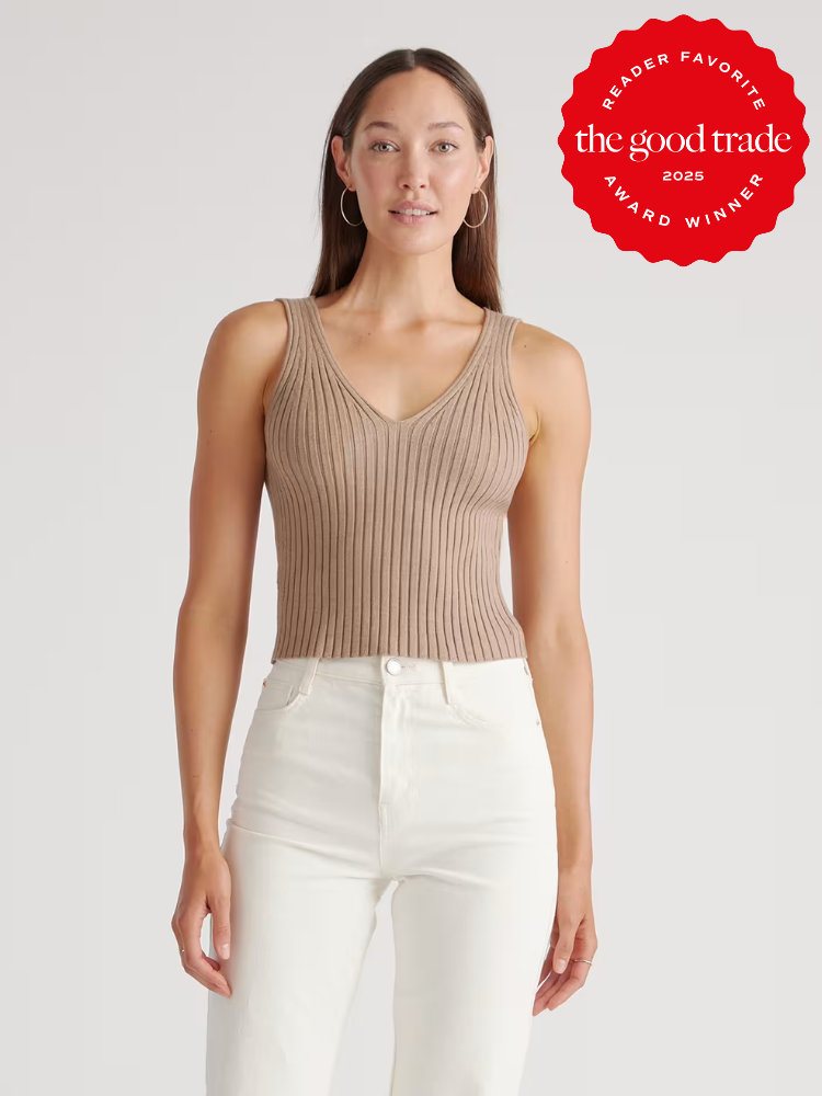 A woman wearing a sleeveless, ribbed beige knit top and high-waisted white jeans stands against a plain background. A red “The Good Trade Award Winner 2025” badge is shown in the top right corner.