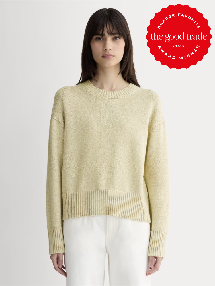 Woman wearing a light yellow knit sweater and white pants stands against a plain background; a red award badge from The Good Trade is displayed in the top right corner.