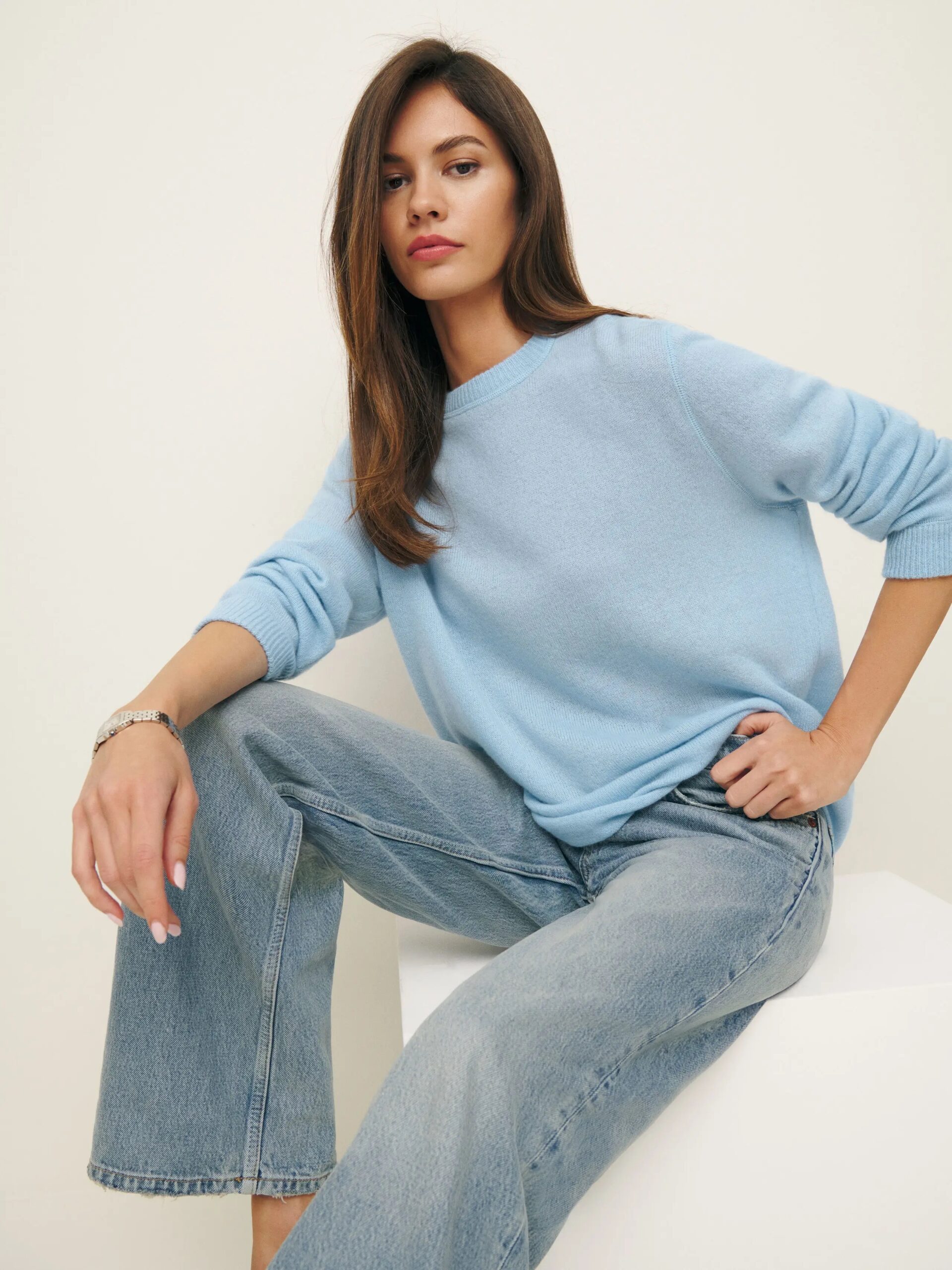 A woman with long brown hair wears a light blue sweater and light blue jeans, sitting on a white surface against a plain background.