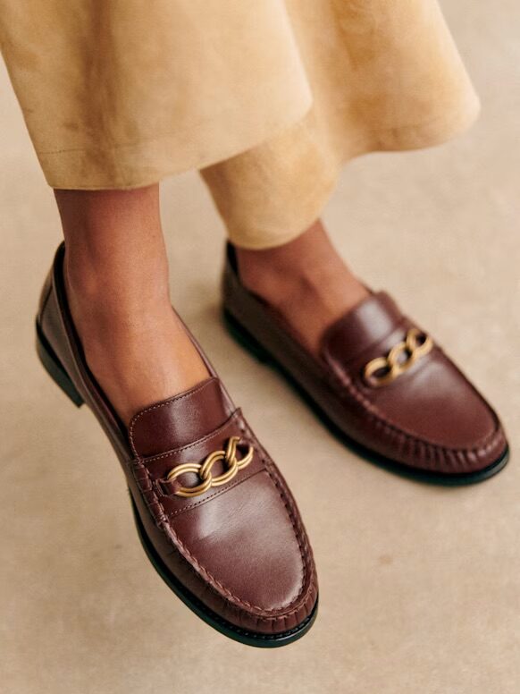 Brown leather loafers with gold chain accents worn with a beige skirt, shown on a person standing on a light-colored floor.