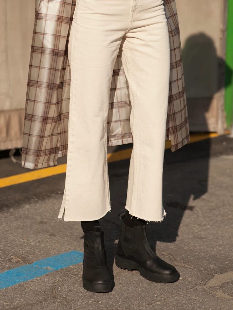 Person wearing cream-colored pants, black boots, and a plaid coat stands on a street with yellow and blue lines. The background includes a gray fabric and green wall.