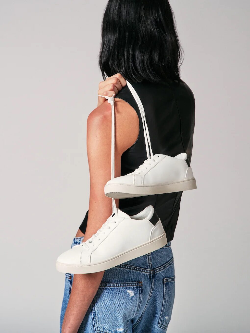Person with long hair wearing a black top and blue jeans holds a pair of white sneakers over their shoulder against a plain background.