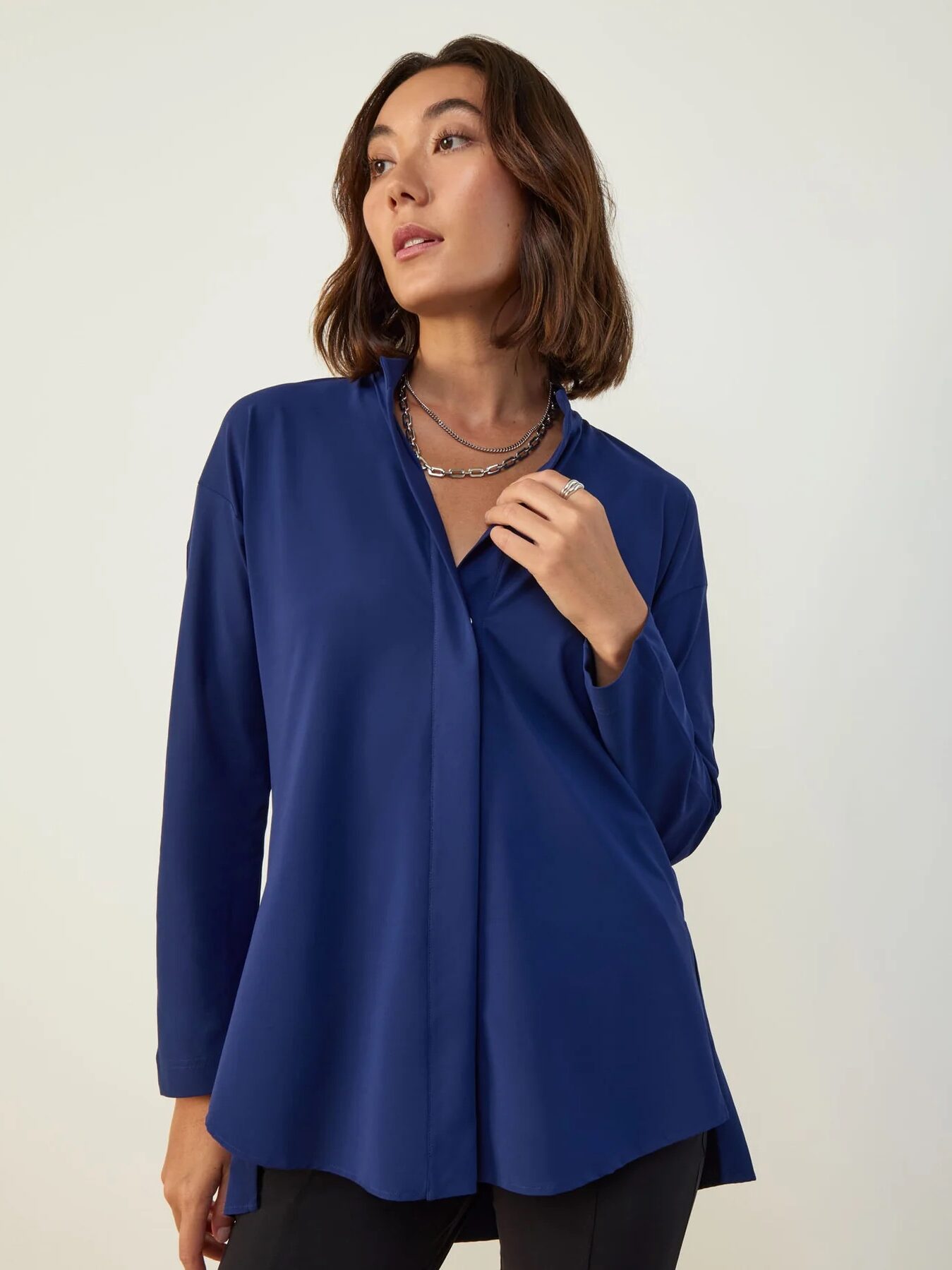 A woman wearing a long-sleeve, deep blue blouse and a silver necklace stands against a plain light background, looking slightly to the side.