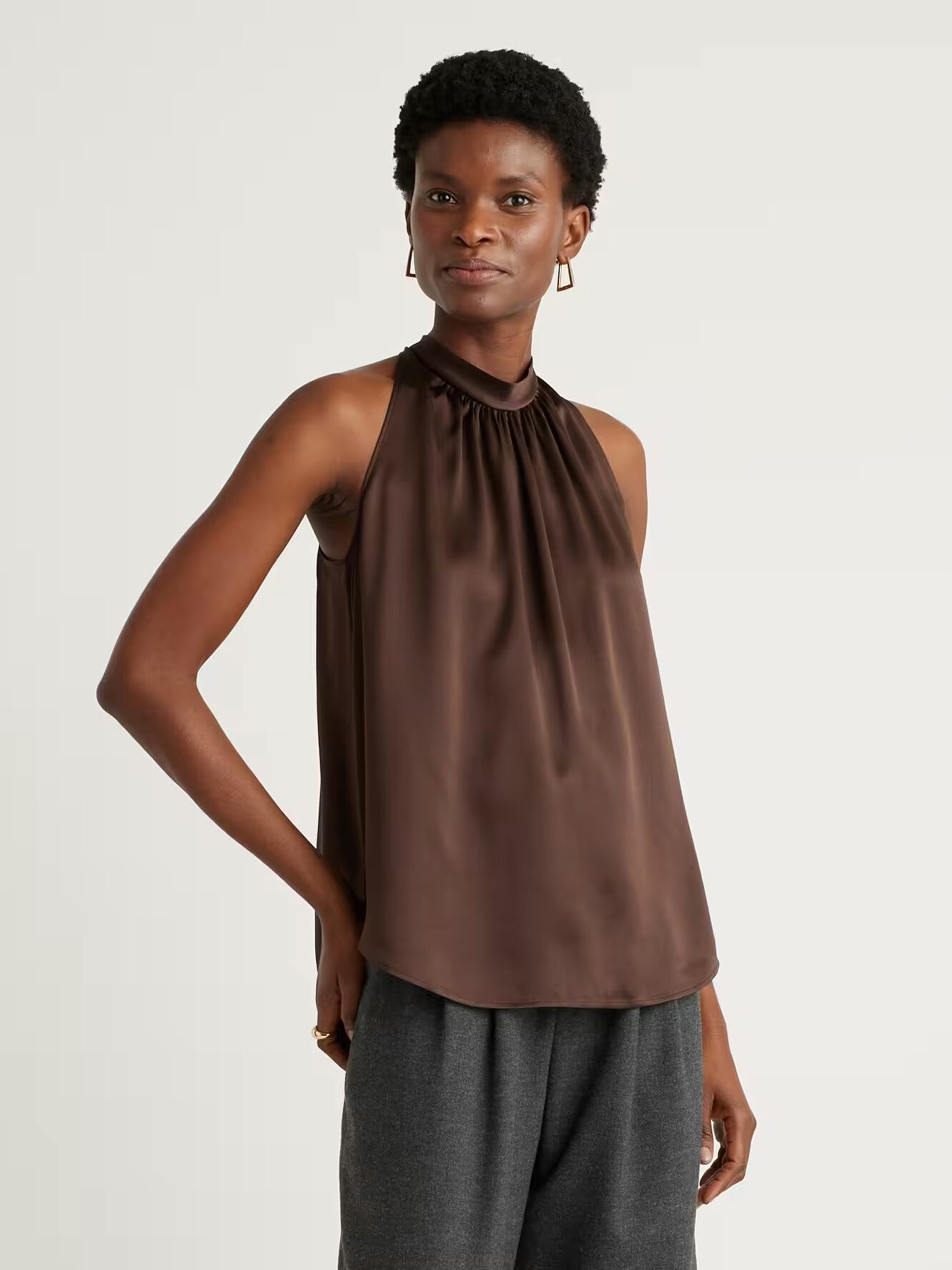 A person stands wearing a sleeveless, brown satin halter top and gray pants against a plain light background.