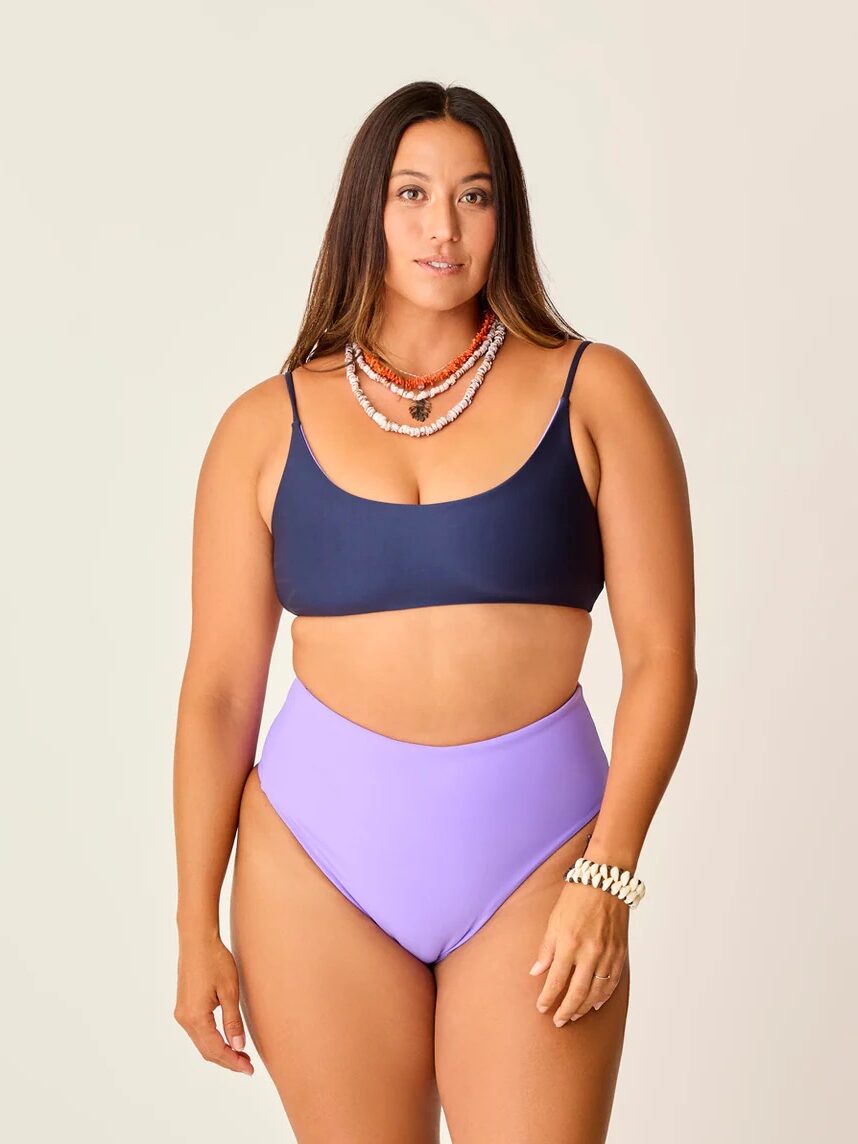 A person wearing a dark bikini top and light purple bottoms stands against a plain background, accessorized with layered necklaces and a bracelet.