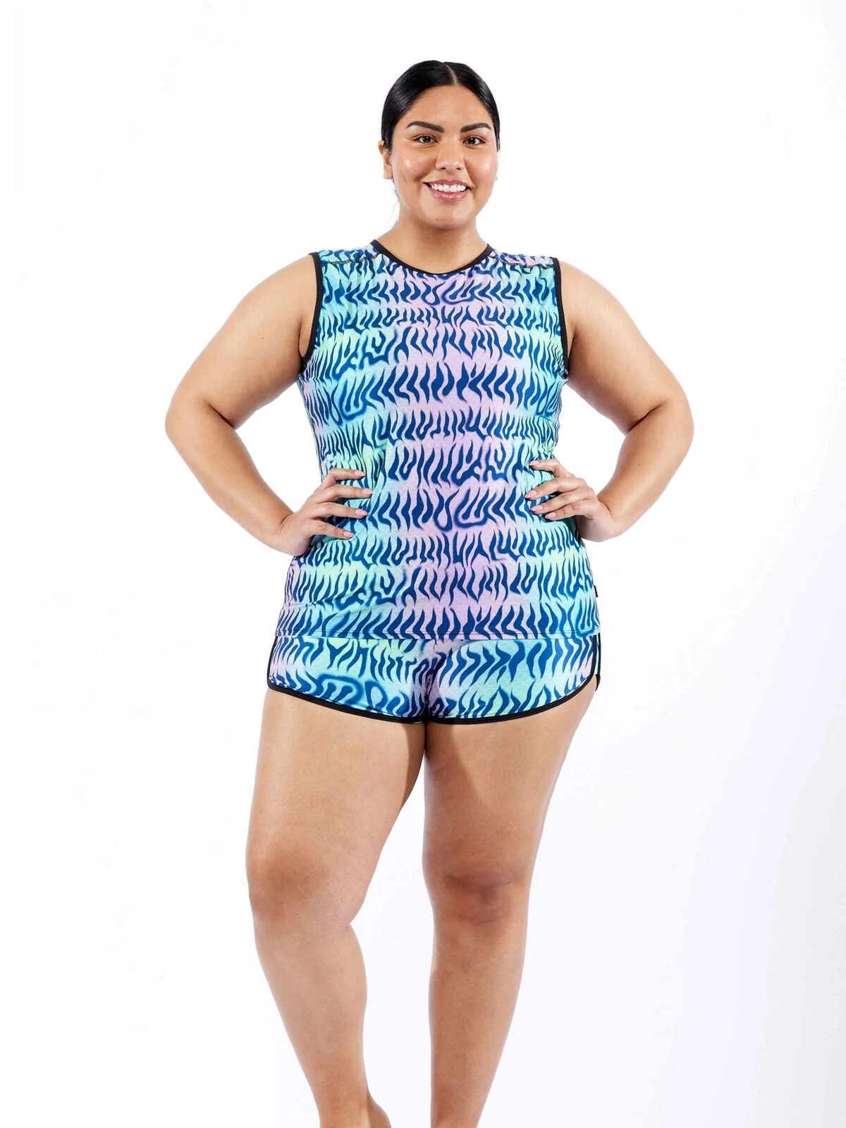 Person wearing a colorful patterned sleeveless romper stands barefoot on a plain white background, with hands on hips and smiling.