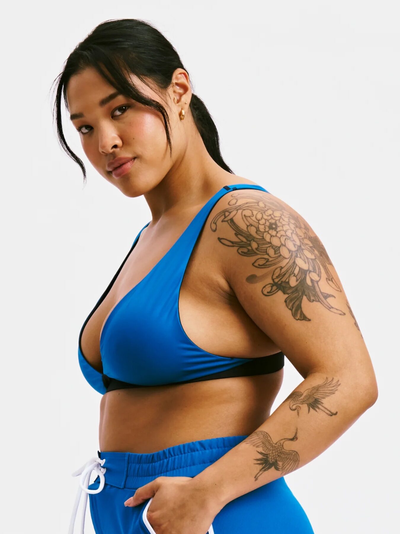 Person standing, wearing a blue sports outfit with visible tattoos on their arm, looking at the camera against a plain white background.
