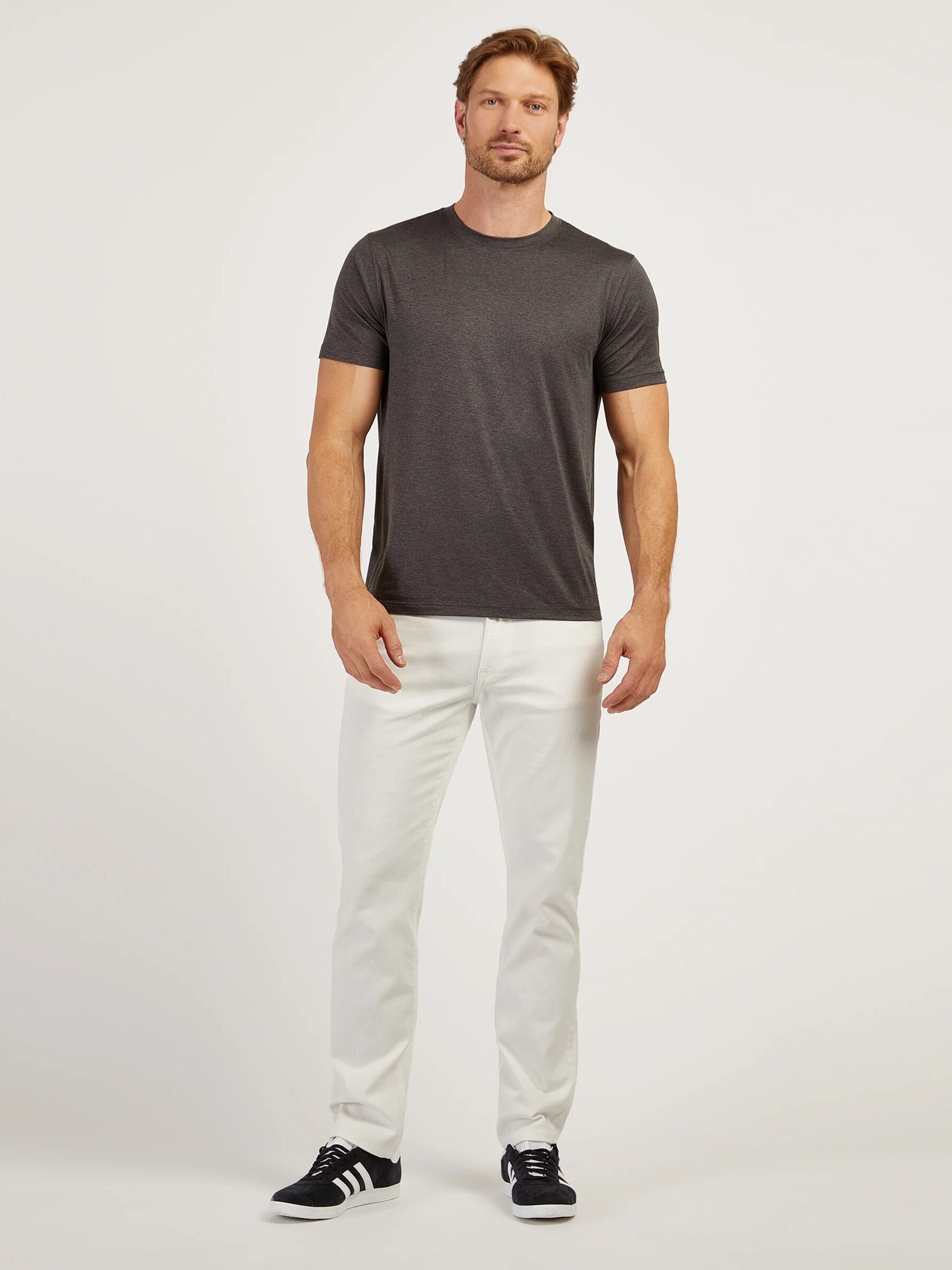 A man stands facing forward wearing a dark gray t-shirt, white pants, and black sneakers against a plain light background.