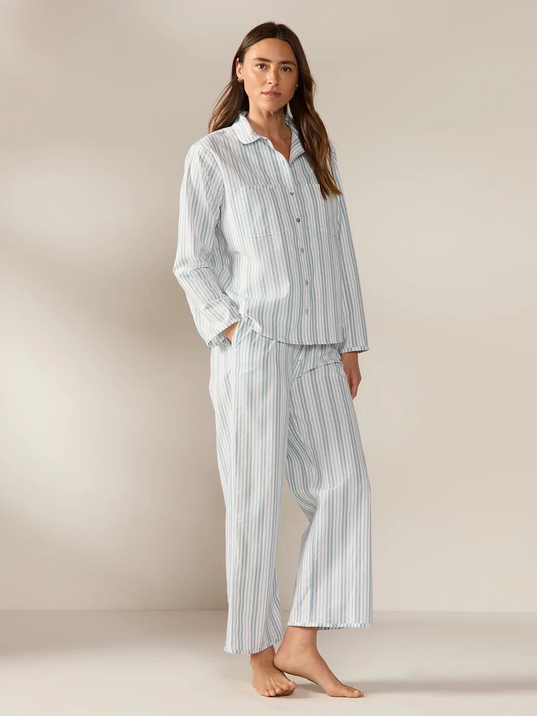A woman stands barefoot on a plain floor, wearing a loose-fitting, long-sleeve, button-up striped pajama set with wide-leg pants.