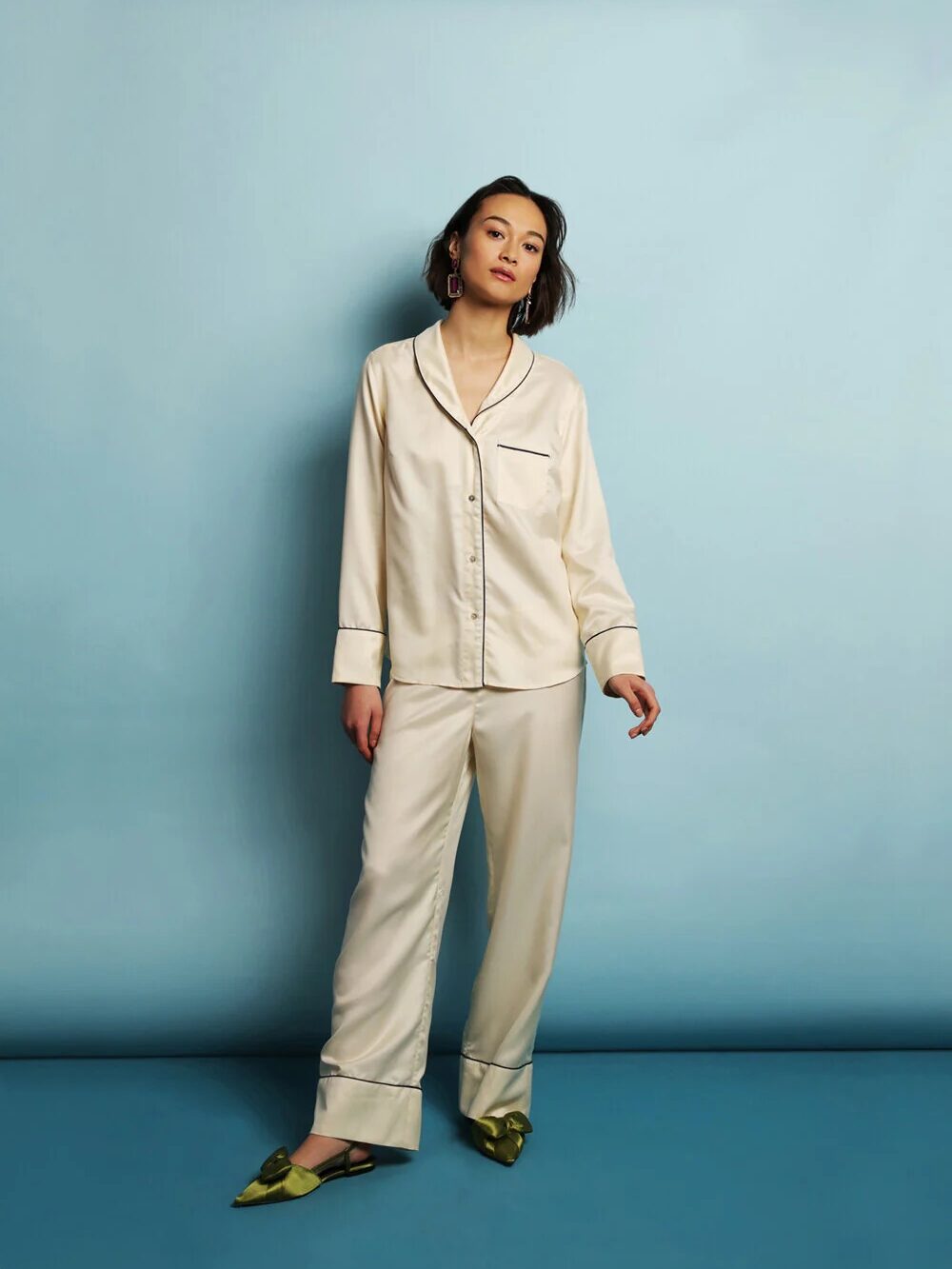 A woman stands against a pale blue background wearing cream-colored satin pajamas and green pointed-toe shoes.