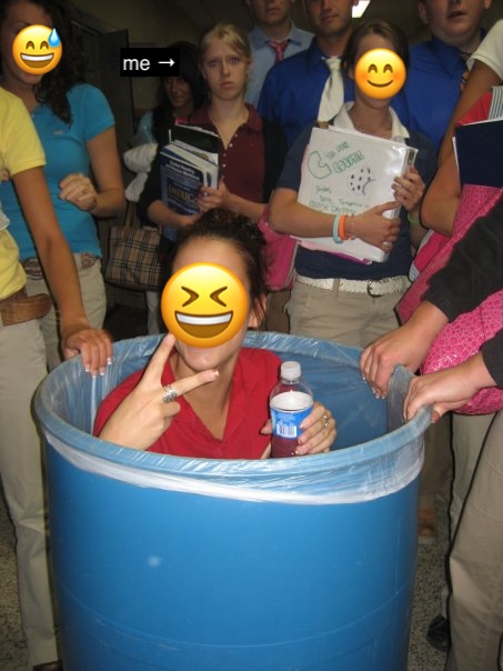 A person sits inside a large blue barrel holding a water bottle and flashing a peace sign, surrounded by students in school uniforms; faces are covered with smiley emojis.