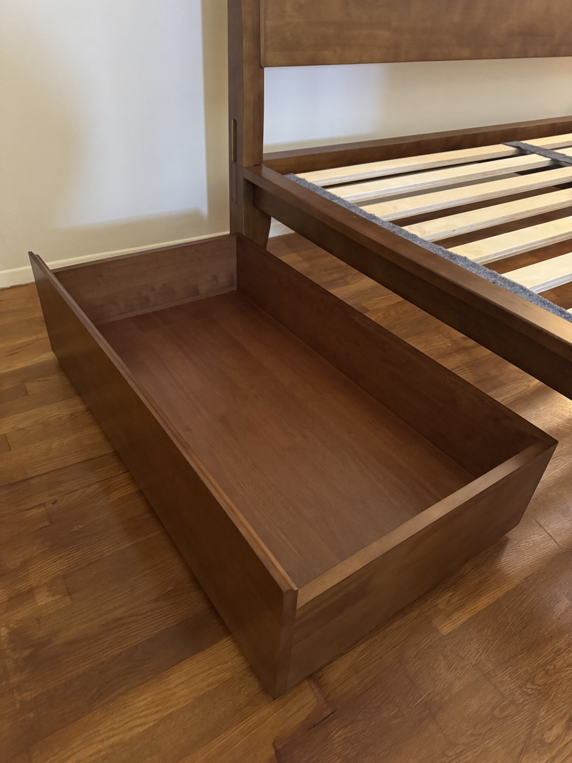 Wooden bed frame with an integrated drawer partially pulled out, on a hardwood floor.