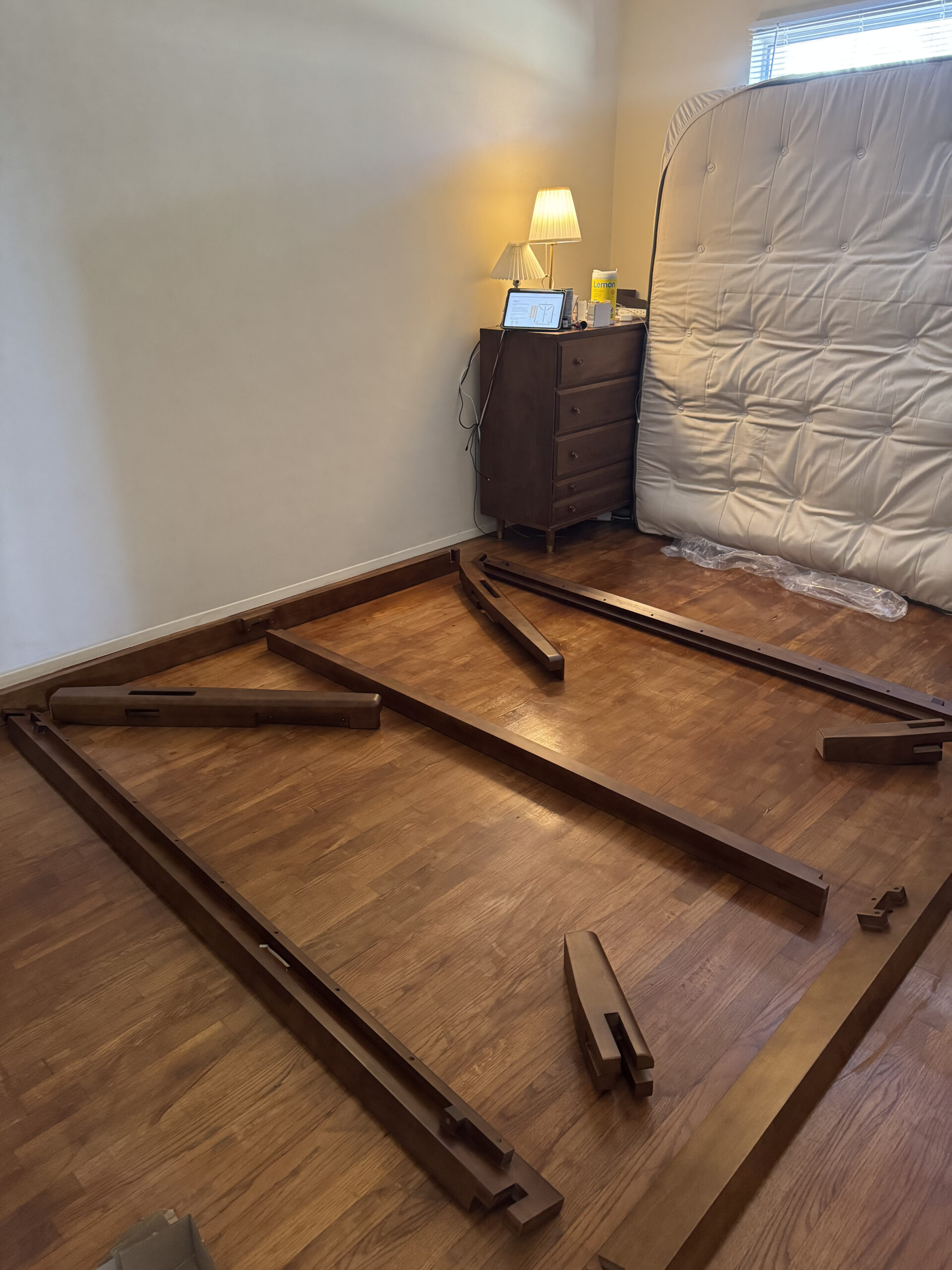 Disassembled bed frame parts on a wooden floor next to a mattress, dresser, lamp, and nightstand in a dimly lit room.