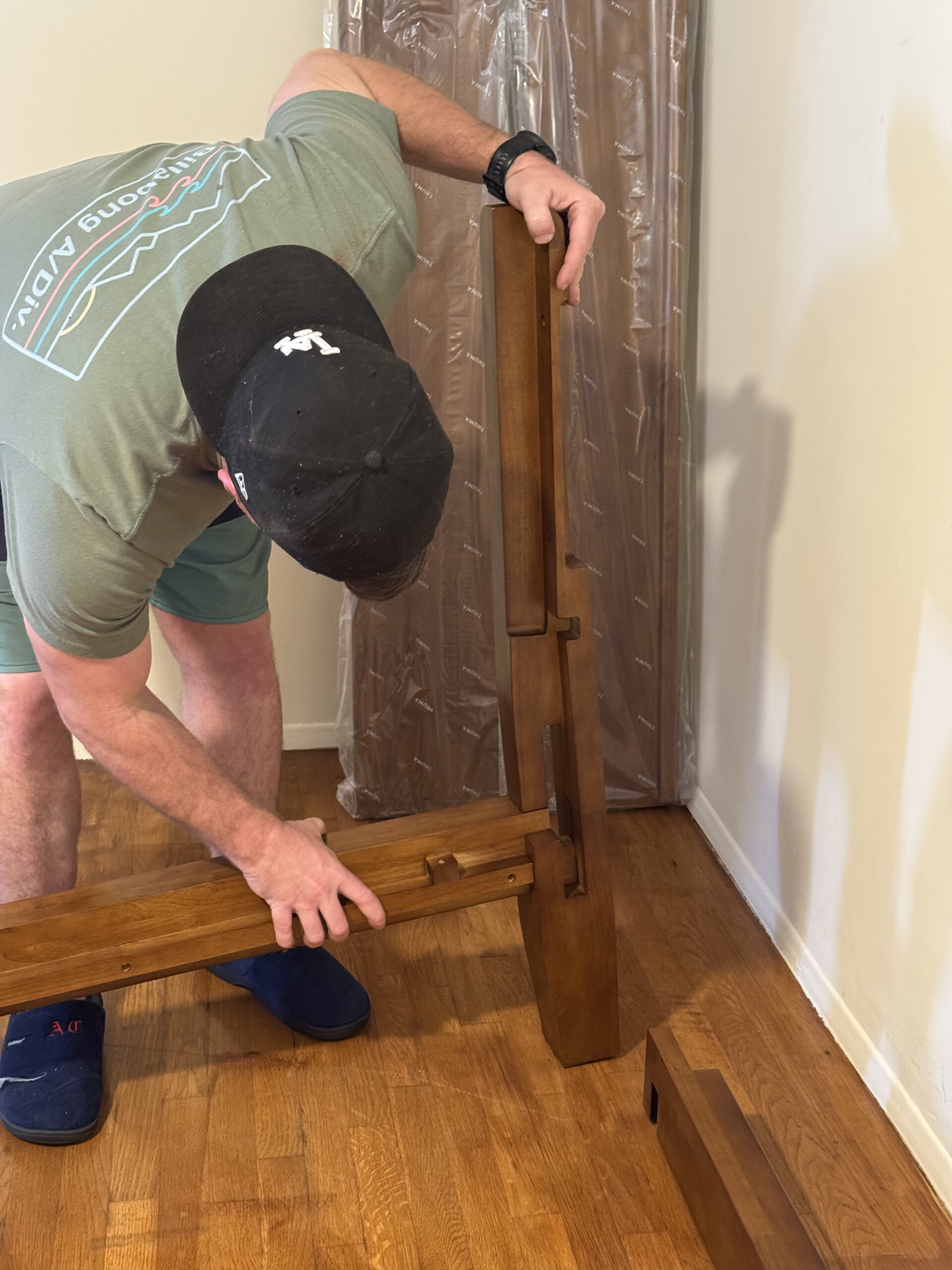Person wearing a cap assembling wooden furniture pieces on a hardwood floor.
