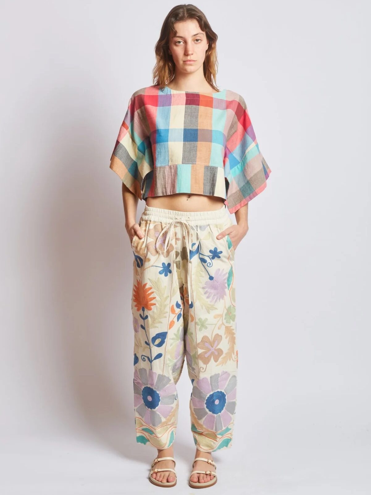 A person stands facing forward, wearing a multicolored plaid crop top, floral print drawstring pants, and sandals against a plain white background.