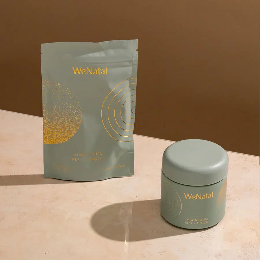 Two WeNatal branded magnesium supplement containers, one in a resealable pouch and the other in a round jar, displayed on a beige surface against a brown background.