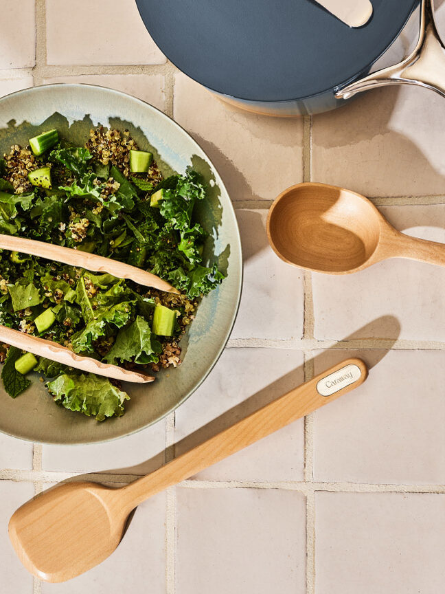 A bowl of salad with leafy greens and cucumber, wooden tongs on top, two wooden utensils, two glasses with lemon water, and napkins on a tiled surface.