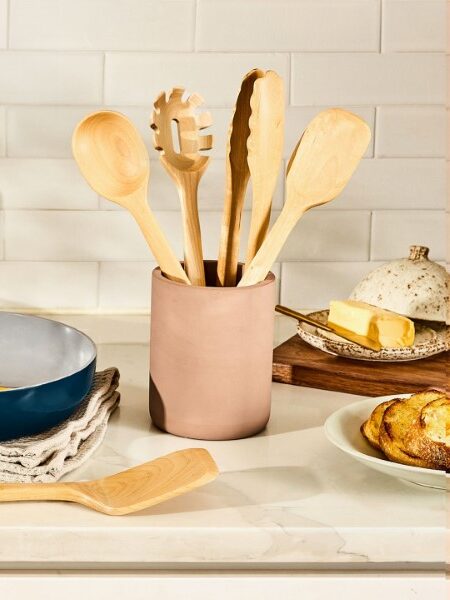 A frying pan with food sits on a towel next to wooden utensils in a container, a pepper grinder, eggs, butter dish, and bread on a kitchen counter.