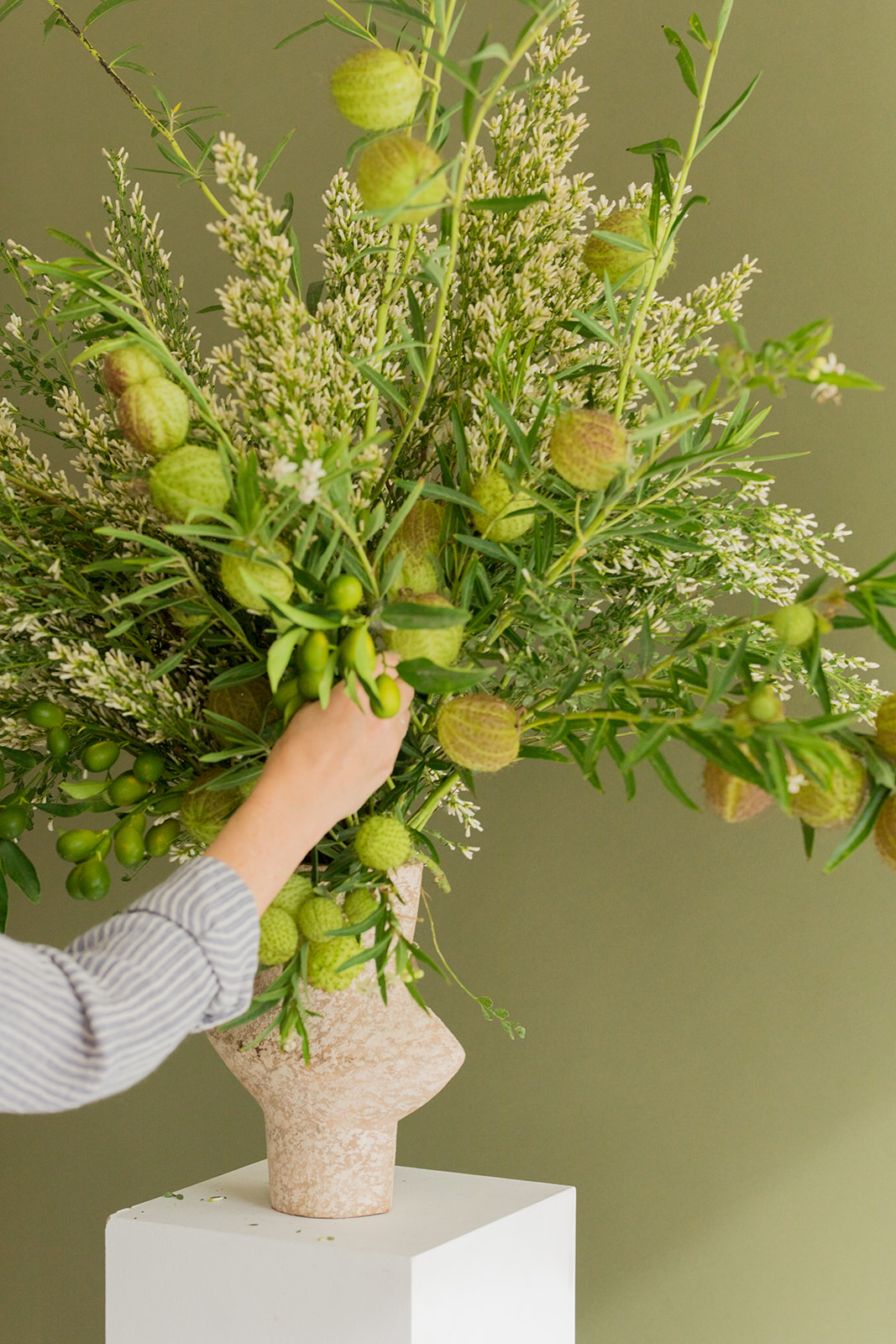A person arranges green foliage and seed pods in a textured beige vase shaped like a head, set against a plain olive green background.