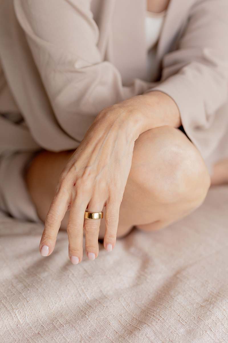 A person in neutral-colored clothing sits with one knee up, resting a hand with a gold ring on their knee. The background is soft and beige.