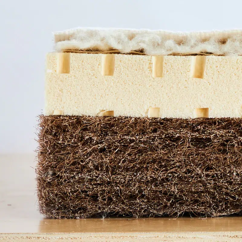 Cross-section of a layered mattress showing a wool top layer, a perforated foam middle layer, and a fibrous brown base layer, all placed on a wooden surface.
