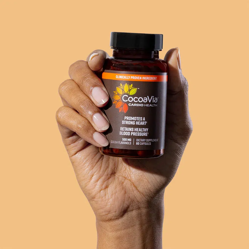 A hand holds a bottle of CocoaVia Cardio Health capsules against a plain light orange background.