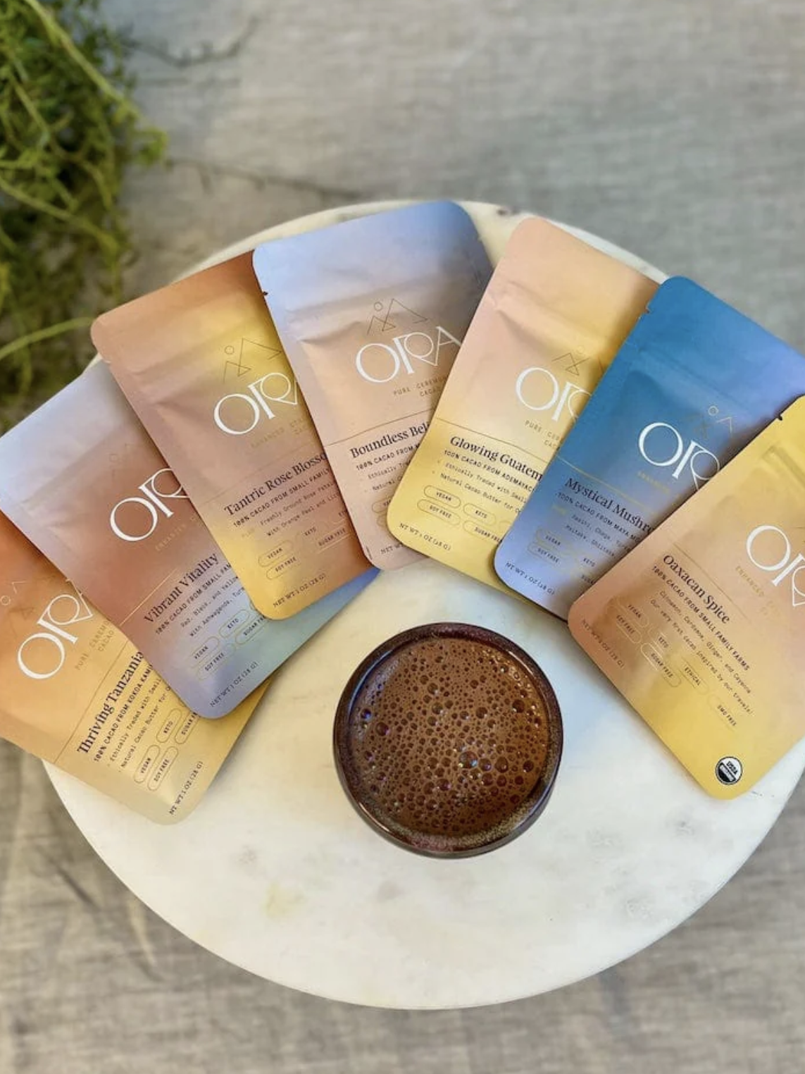 A round table displays a cup of frothy coffee surrounded by six colorful ORA coffee packets arranged in a semicircle.