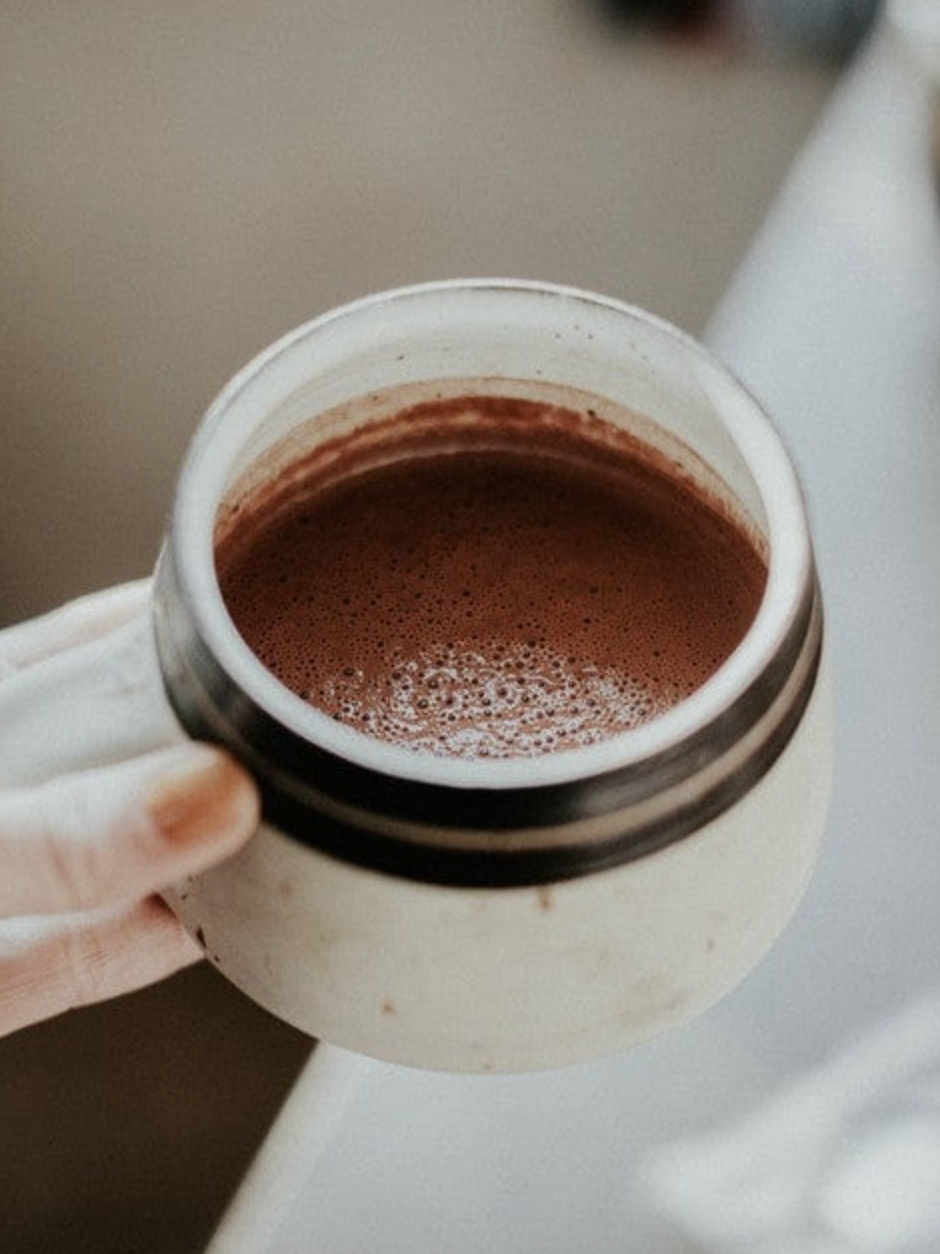 A hand holding a white ceramic mug filled with hot chocolate or coffee, with a visible ring on one finger and a blurred background.