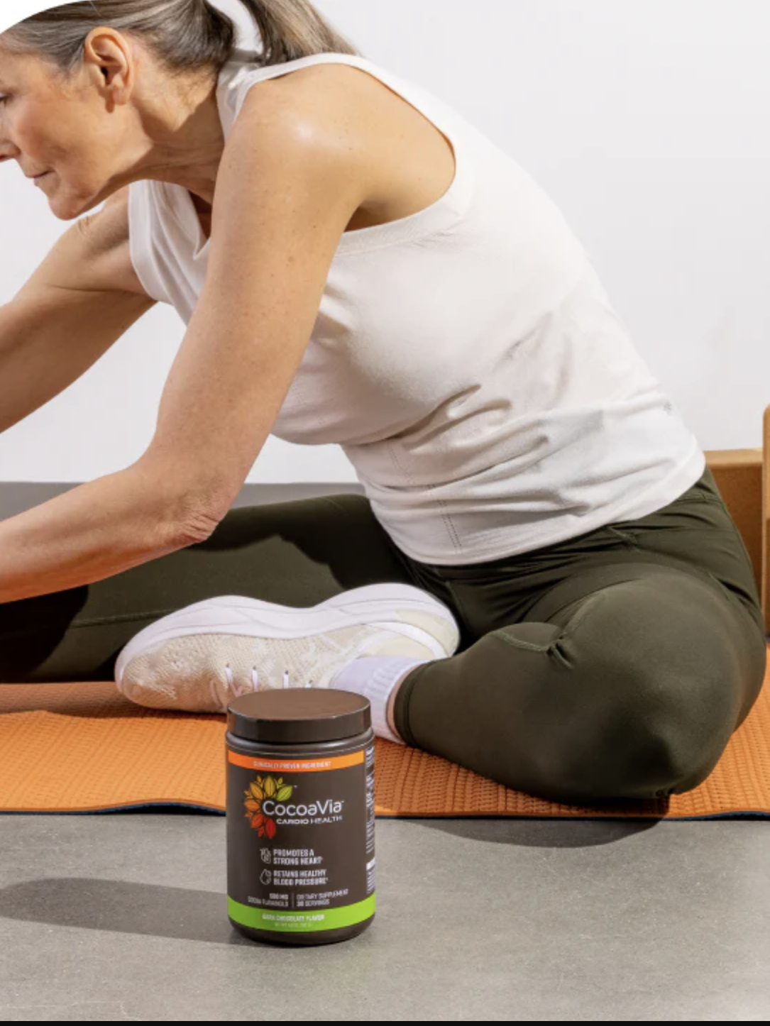 A woman stretches on an orange yoga mat next to a jar of CocoaVia supplement, a green kettlebell, yoga blocks, and a water bottle.