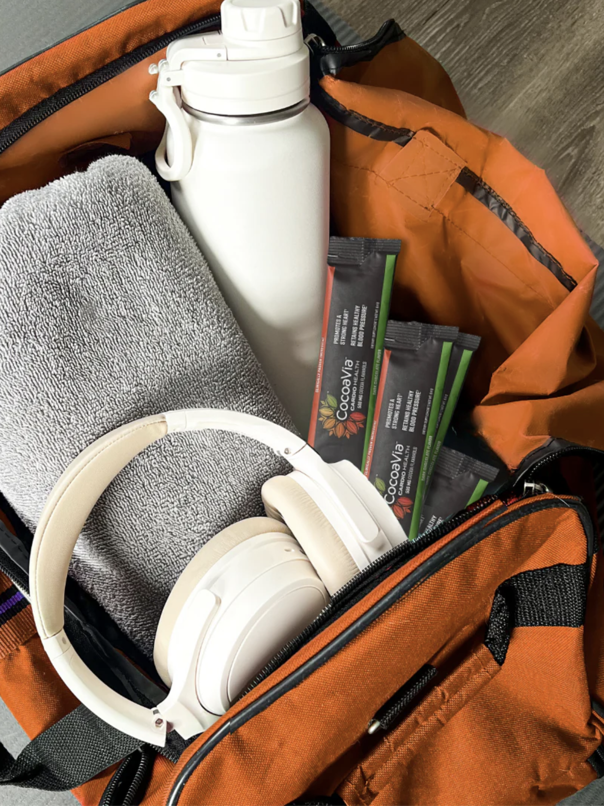 An open orange gym bag containing a gray towel, a white water bottle, several drink mix packets, and a pair of white headphones.