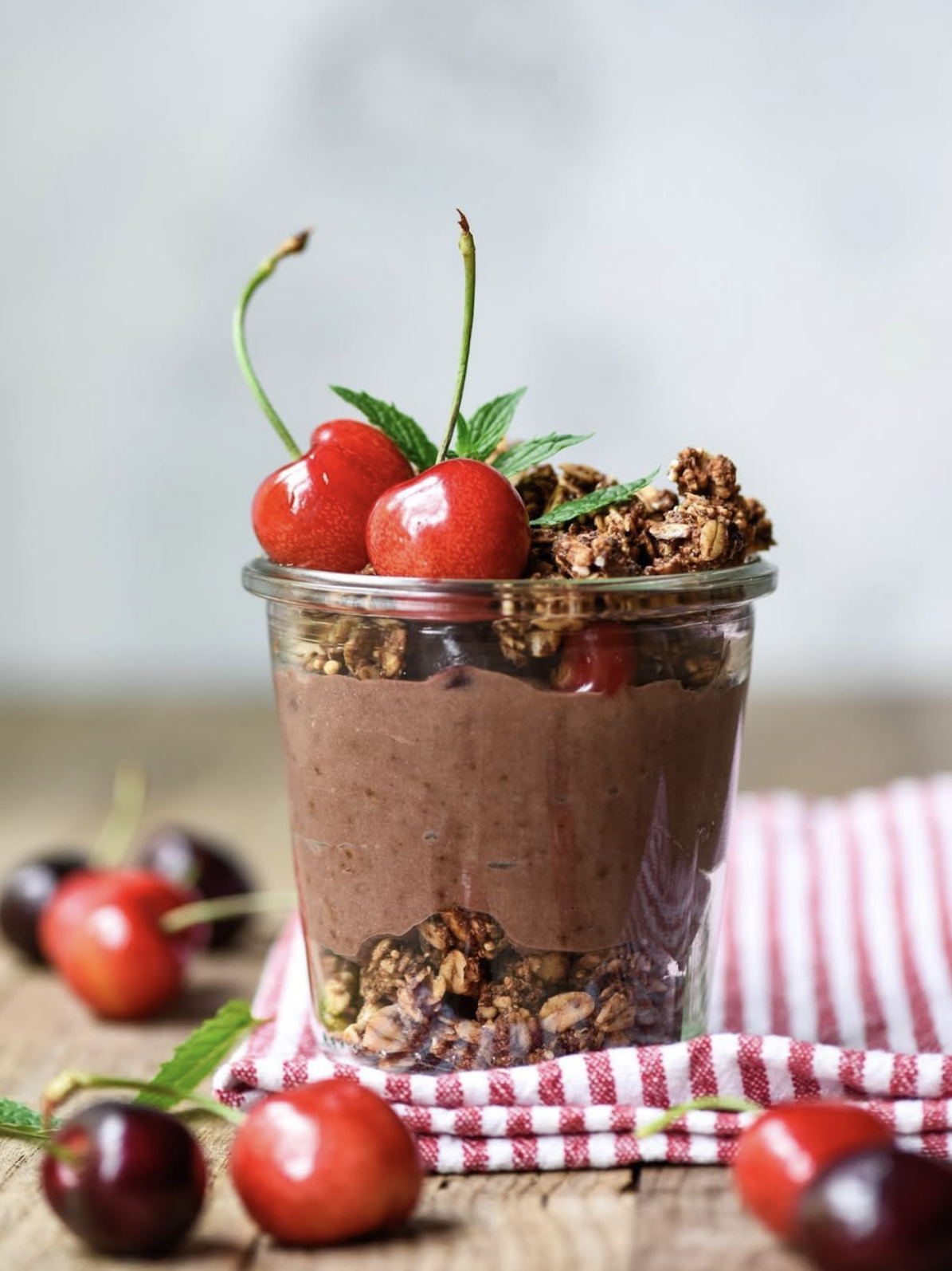 A glass jar filled with chocolate mousse and granola, topped with two fresh cherries and a mint sprig, sits on a striped cloth with cherries scattered around.