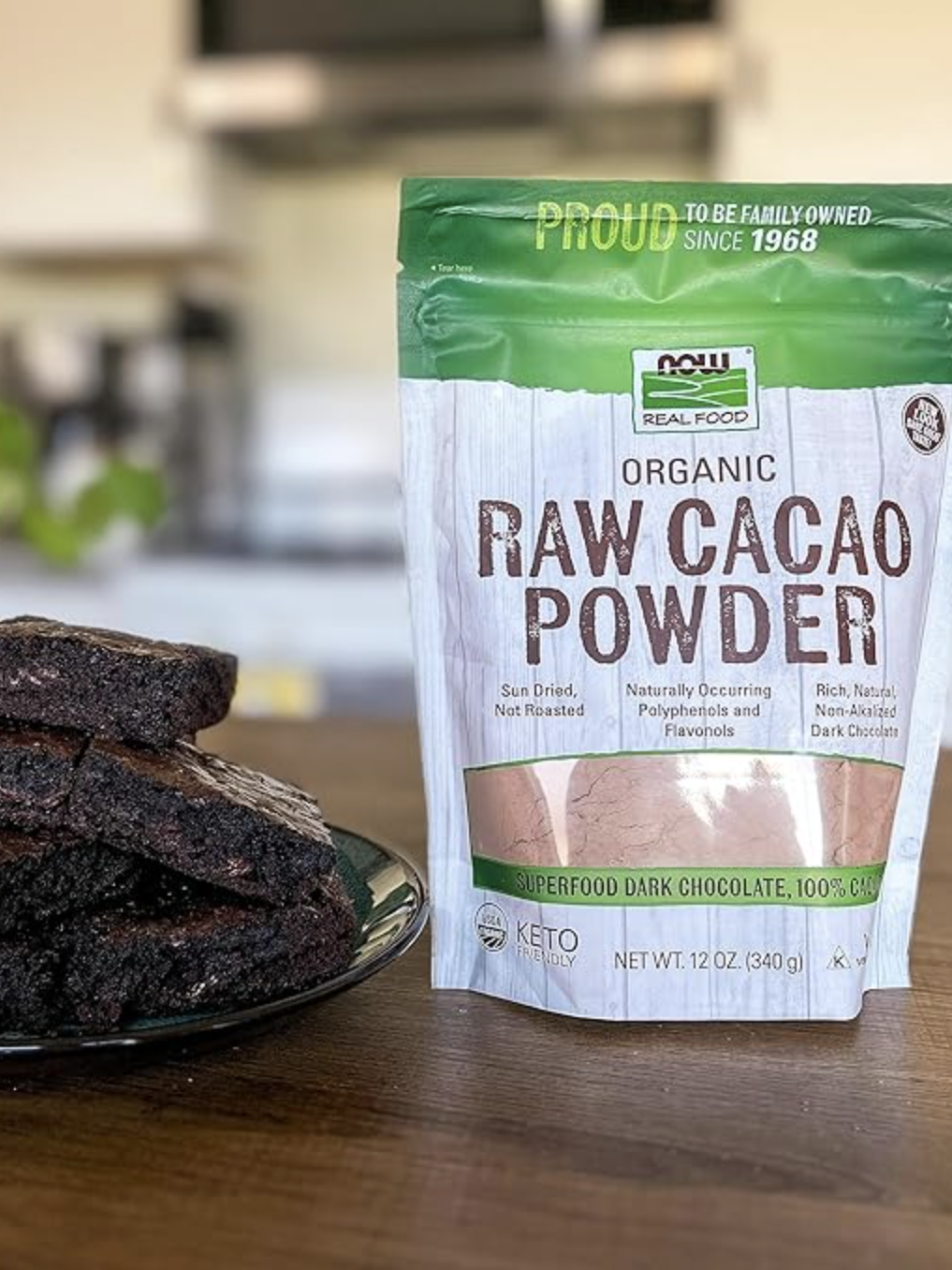 A plate of brownies sits beside a standing bag of NOW Real Food Organic Raw Cacao Powder on a kitchen counter.