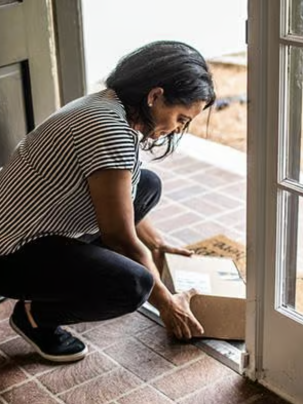 A person in a striped shirt kneels by an open door, picking up a package from the doormat inside the entryway.