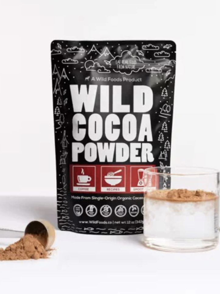A bag of Wild Cocoa Powder stands upright beside a glass cup containing cocoa powder and a metal scoop with cocoa powder spilled on a white surface.