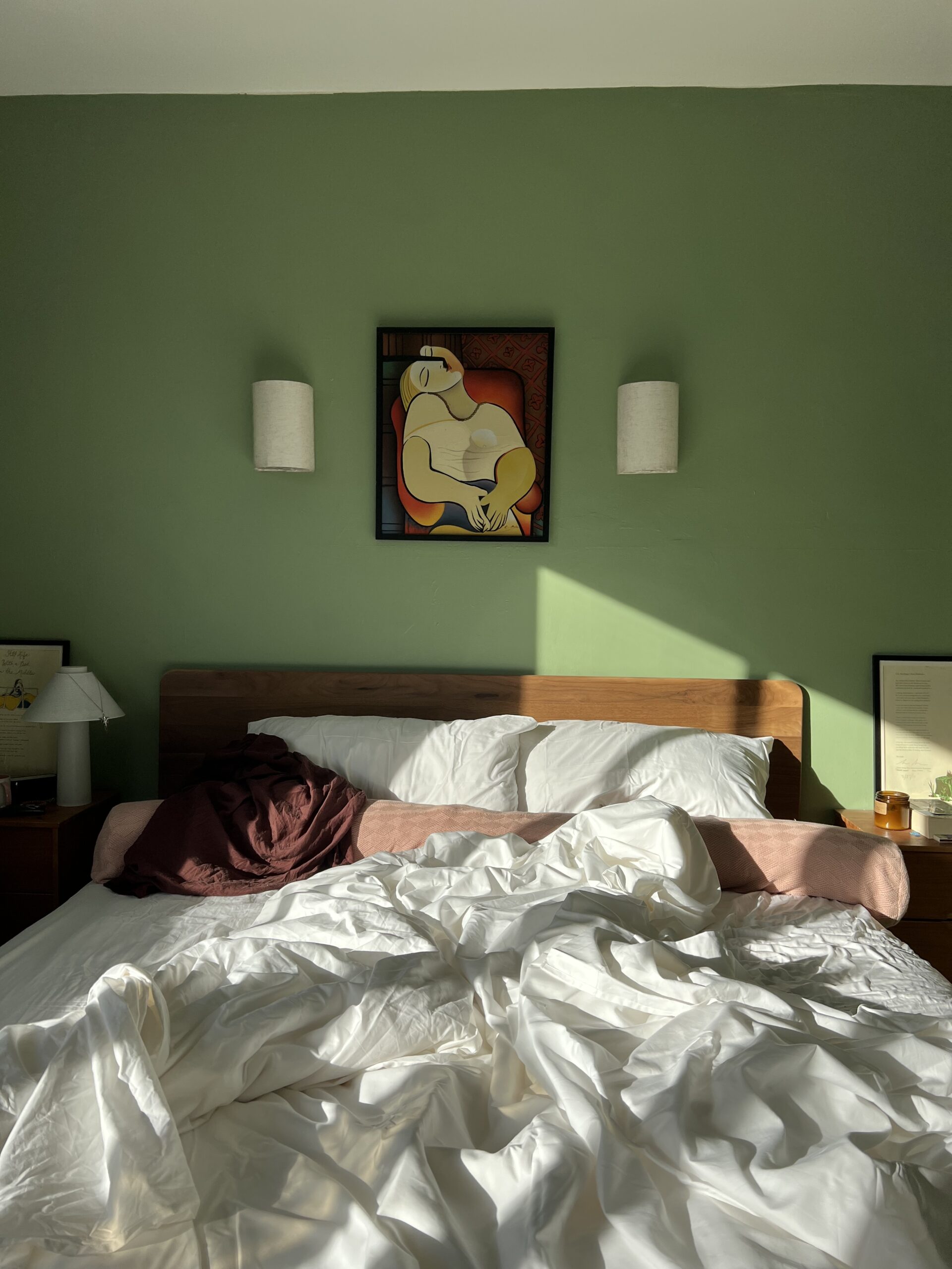 Unmade bed with white bedding and a brown blanket, beneath a painting of an abstract figure on a green wall, flanked by two wall lights. Sunlight falls across the scene.