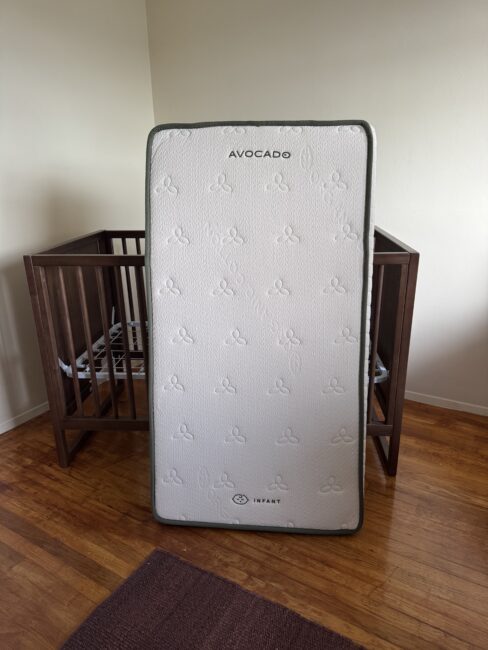 A white Avocado brand infant mattress stands upright in front of a wooden crib in a room with wood flooring.