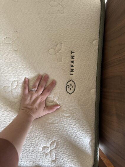 A hand presses down on a crib mattress labeled "INFANT" with a baby face icon, showing the mattress surface and texture.
