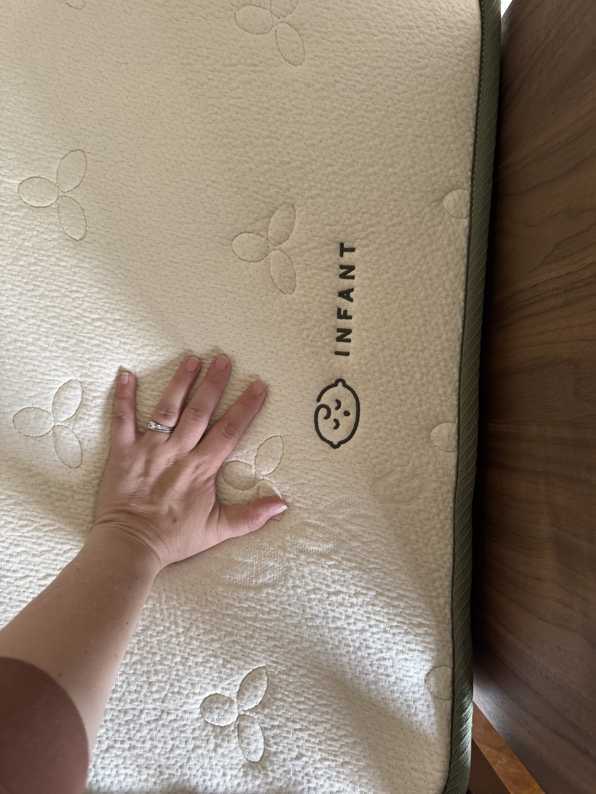 A hand presses down on a crib mattress labeled "INFANT" with a baby face icon, showing the mattress surface and texture.
