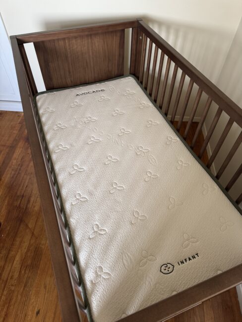 A wooden crib with a white Avocado brand infant mattress inside, placed on a hardwood floor near a beige wall.
