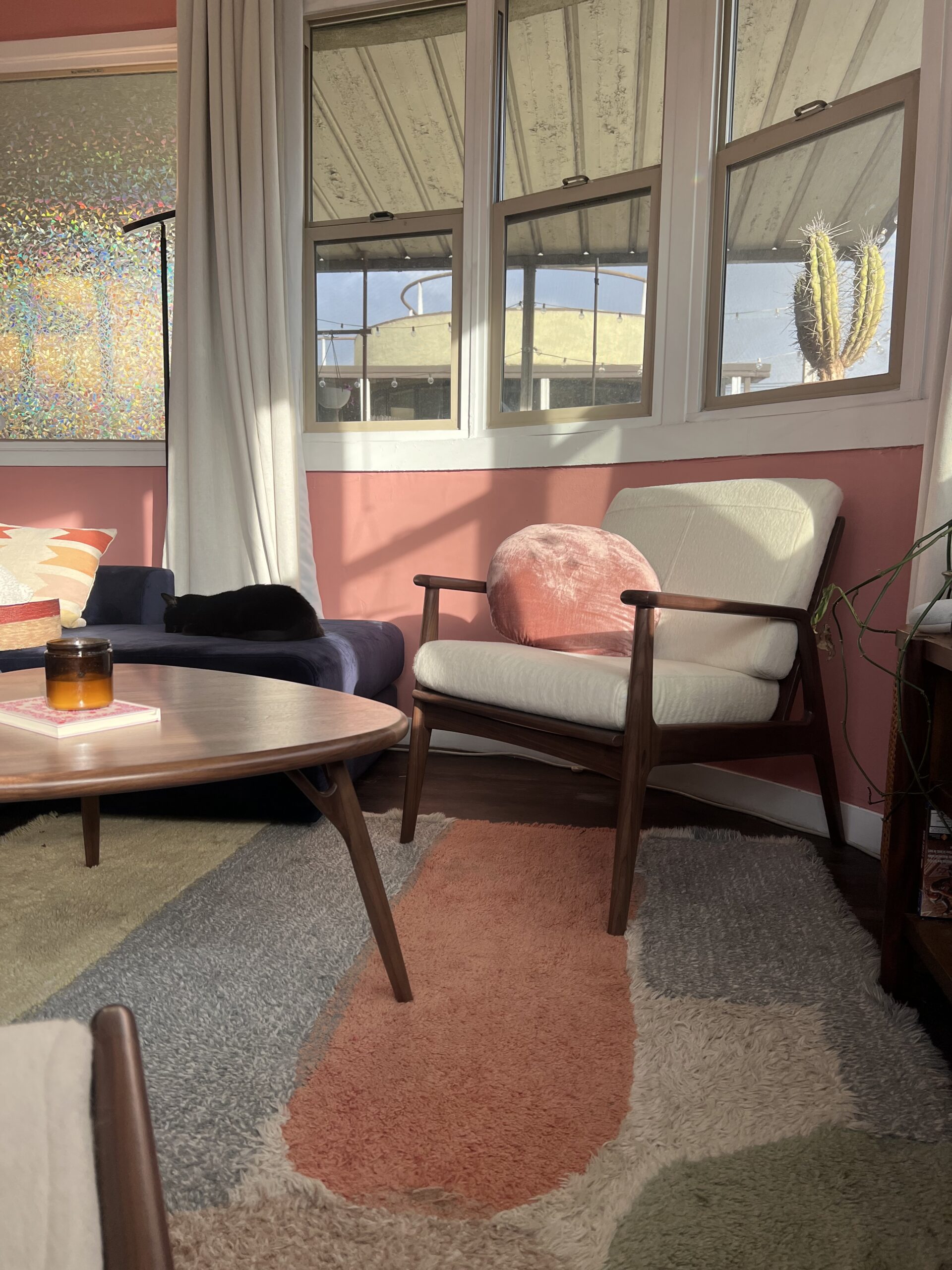 A sunlit living room with a white cushioned chair, a round pillow, a wooden coffee table with a candle and book, a couch, a cactus, and patterned rugs.