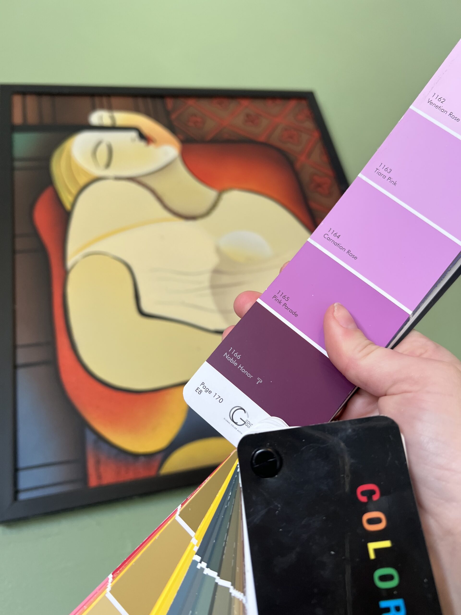 A hand holds a paint color swatch with various shades of pink and purple in front of a framed abstract painting of a reclining woman on a green wall.