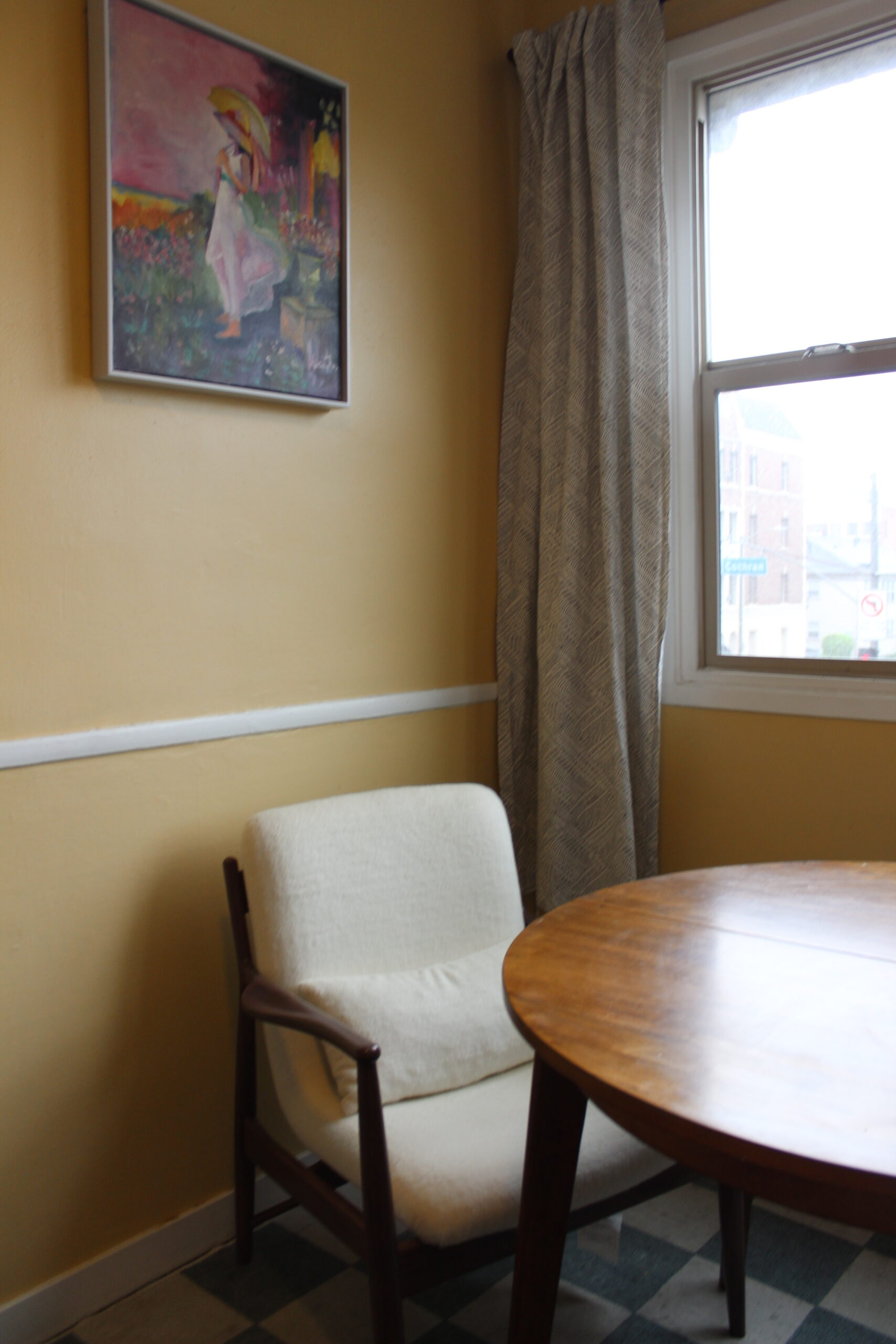 A wooden chair with a white cushion sits next to a round wooden table in a yellow room with a window and a framed painting on the wall.
